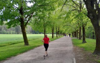 Lenkkeilijä punaisessa paidassa juoksee vehreiden lehtipuiden reunustamaa kesäistä hiekkatietä pitkin.