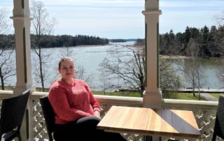 Villa Wolaxin palvelupäällikkö Milla Ahorinta istuu terassilla pöydän ääressä. Taustalla näkyy mereinen saaristomaisema.