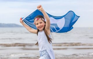 Nuori tyttö vaalenasinisessä mekossa juoksee hiekkarannalla pitäen sinivalkoista Blue flag -lippua pääänsä yläpuolella. Tytöllä on päässään vaaleanpunainen kukkaseppele.