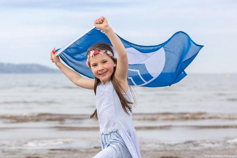 Nuori tyttö vaalenasinisessä mekossa juoksee hiekkarannalla pitäen sinivalkoista Blue flag -lippua pääänsä yläpuolella. Tytöllä on päässään vaaleanpunainen kukkaseppele.