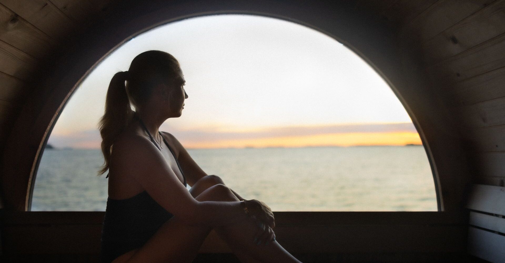 Nainen katselee vesistöä puolipyöreästä ikkunasta hämärässä saunassa.