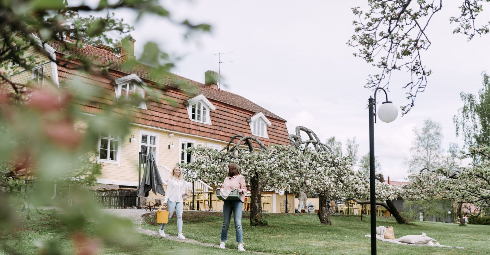 Taustalla kaunis puinen rakennus, keväiset omenapuut kukkivat, kaksi naista kävelee talon edessä polkua pitkin.