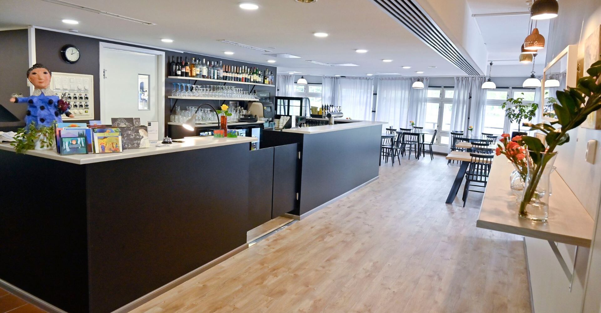 Kuvassa on Koto hotellin vastaanottotiski valoisassa huoneessa. Tiskillä seisoo iso kangasnukke sinisessä puvussa, ottamassa vieraita vastaan.