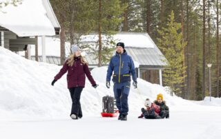 Äiti ja isä kävelevät lumen peittämien tupien edustalla vetäen perässään kahta pulkkaa. Toisessa pulkassa istuu kaksi pientä lasta ja toisessa vedetään matkalaukkua.