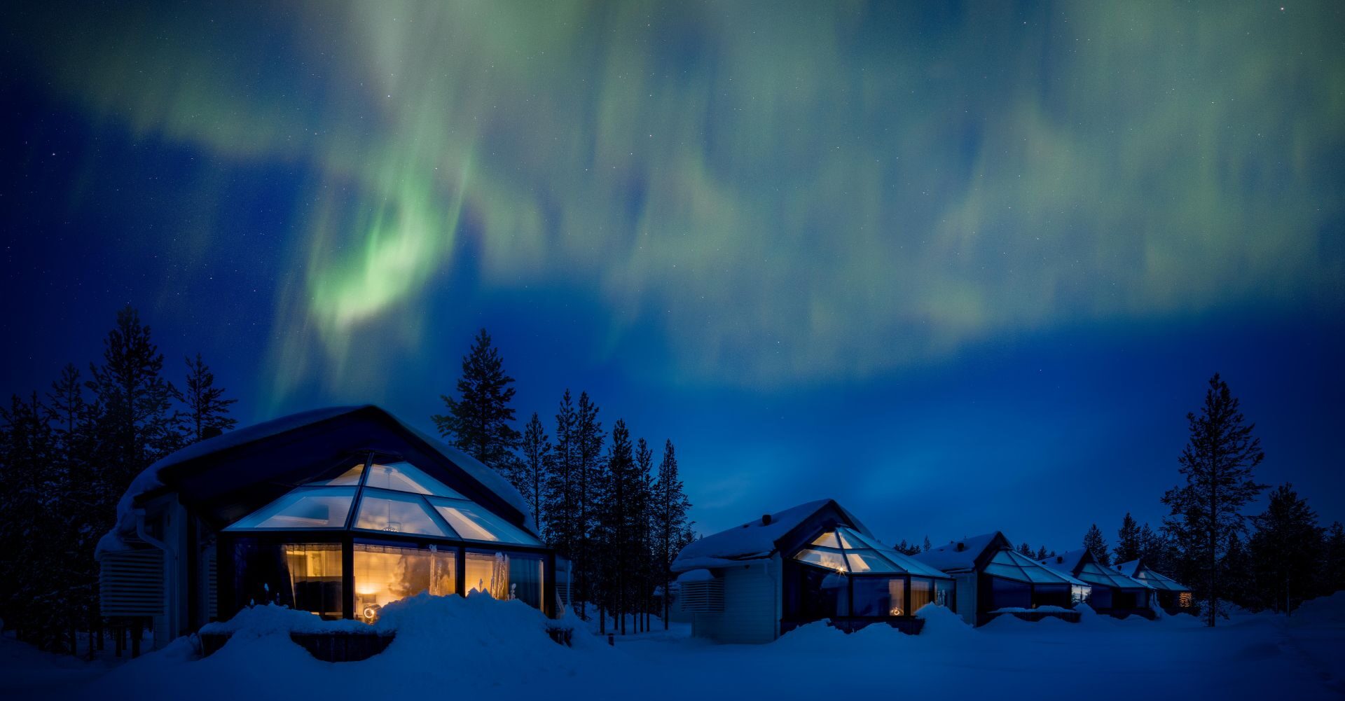 Kolme majoitusiglua seisoo lumisessa talvimaisemassa vihreiden rvontulien valaiseman yötaivaan alla.
