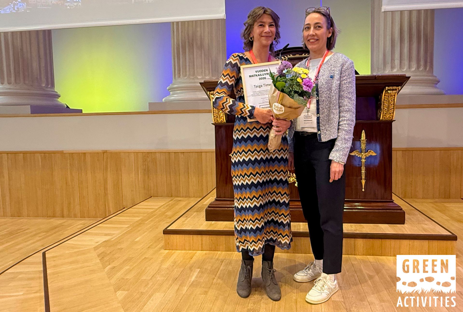 Kuvassa Taiga Timesin Kiia poseeraa Vuoden matkailuyritys -diplomin ja kukkakimpun kera, vierellään Going Greenin ohjelmajohtaja Heidi Heinonen.