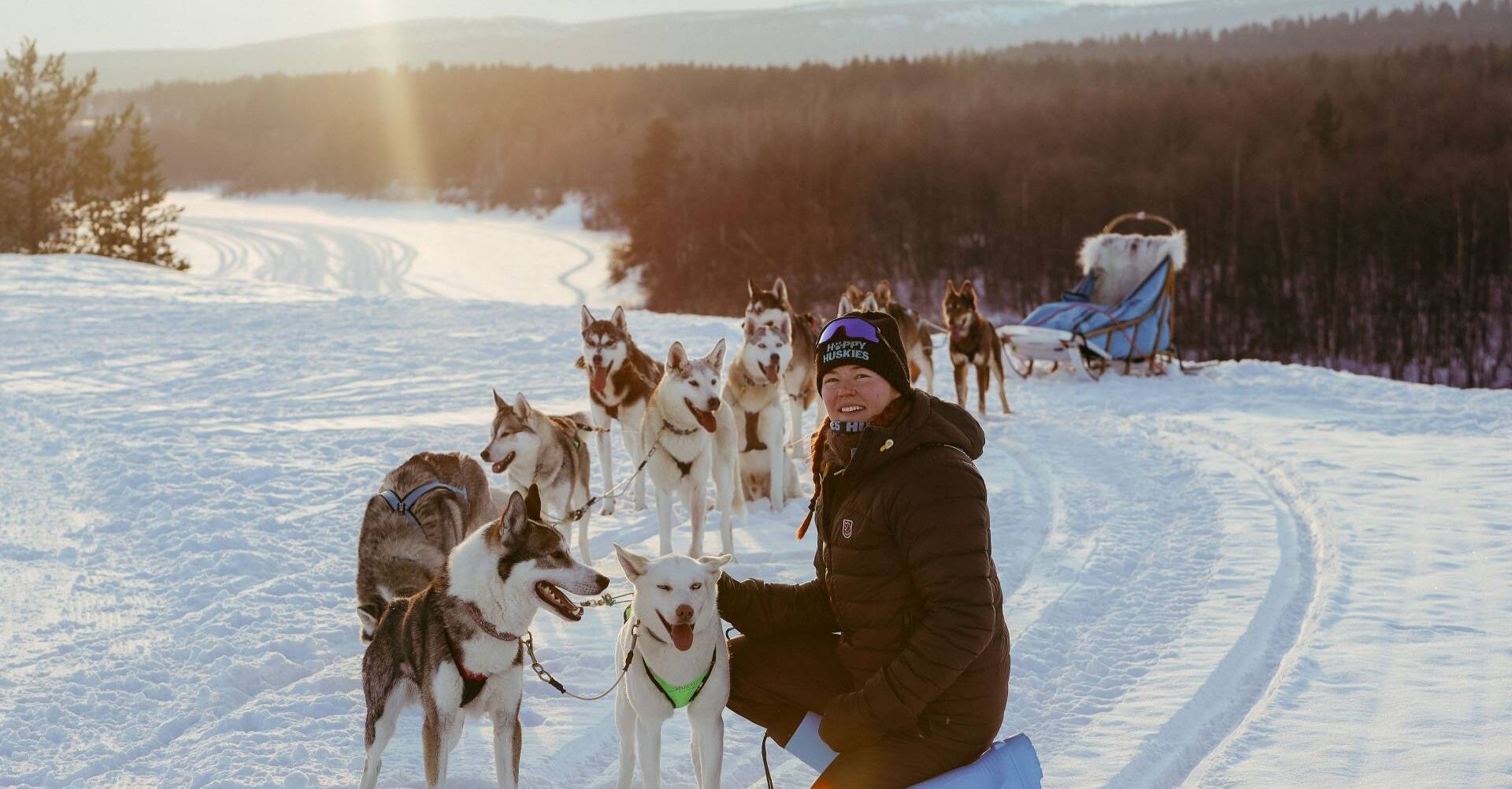 Kuvassa on koiravaljakko tunturimaisemassa. Edustalla istuu hymyilevä nainen, joka rapsuttaa etummaisen koiran päätä, koiralla on kieli ulkona ja hän on tyytyväisen näköinen. Aurinko pilkottaa pilvien lomasta.