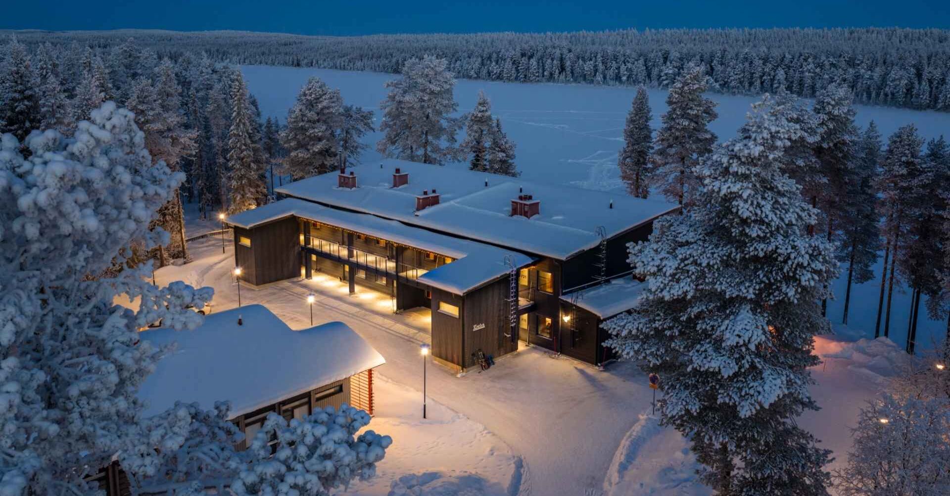 Talvinen kuva yläilmoista Oivangin pimeässä valaistuta päärakennuksesta, jonka taustalla näkyy lumen peittämä vesistö ja sen takana tiheä kuusimetsä.