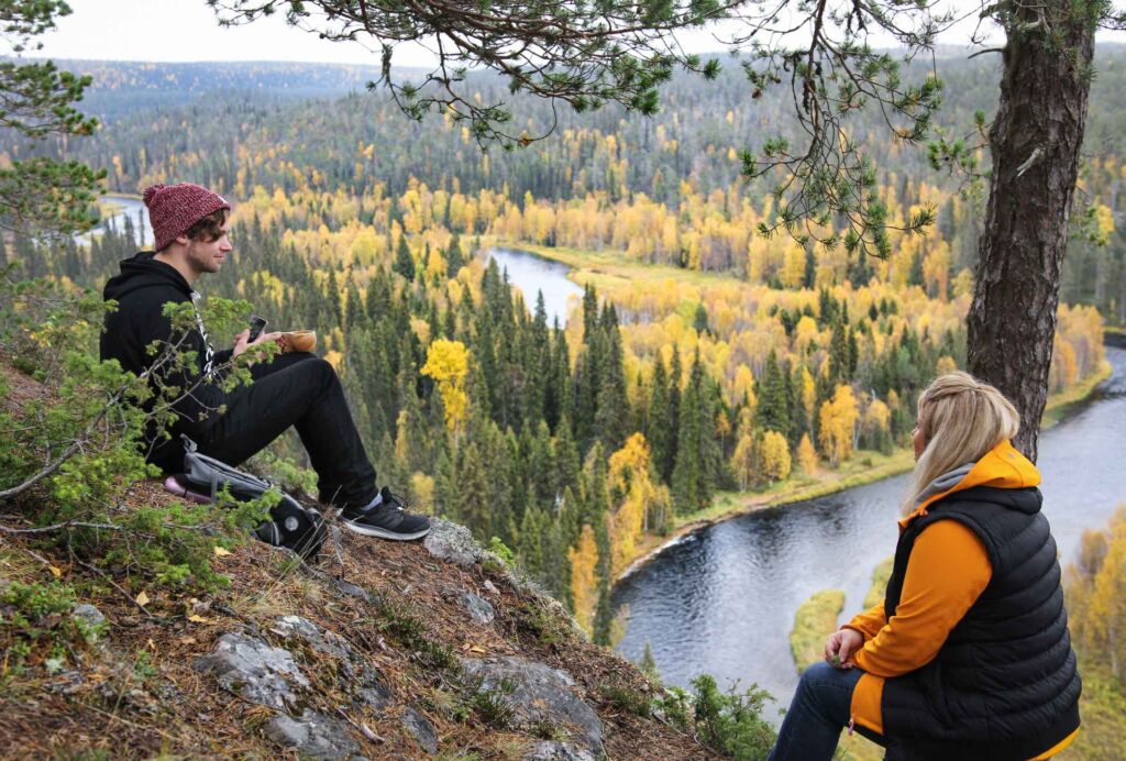 Kalliolla istuu nainen ja mies. Alhaalla avautuu kaunis maisema, jossa näkyy ruskan värjäämiä puita ja joki.